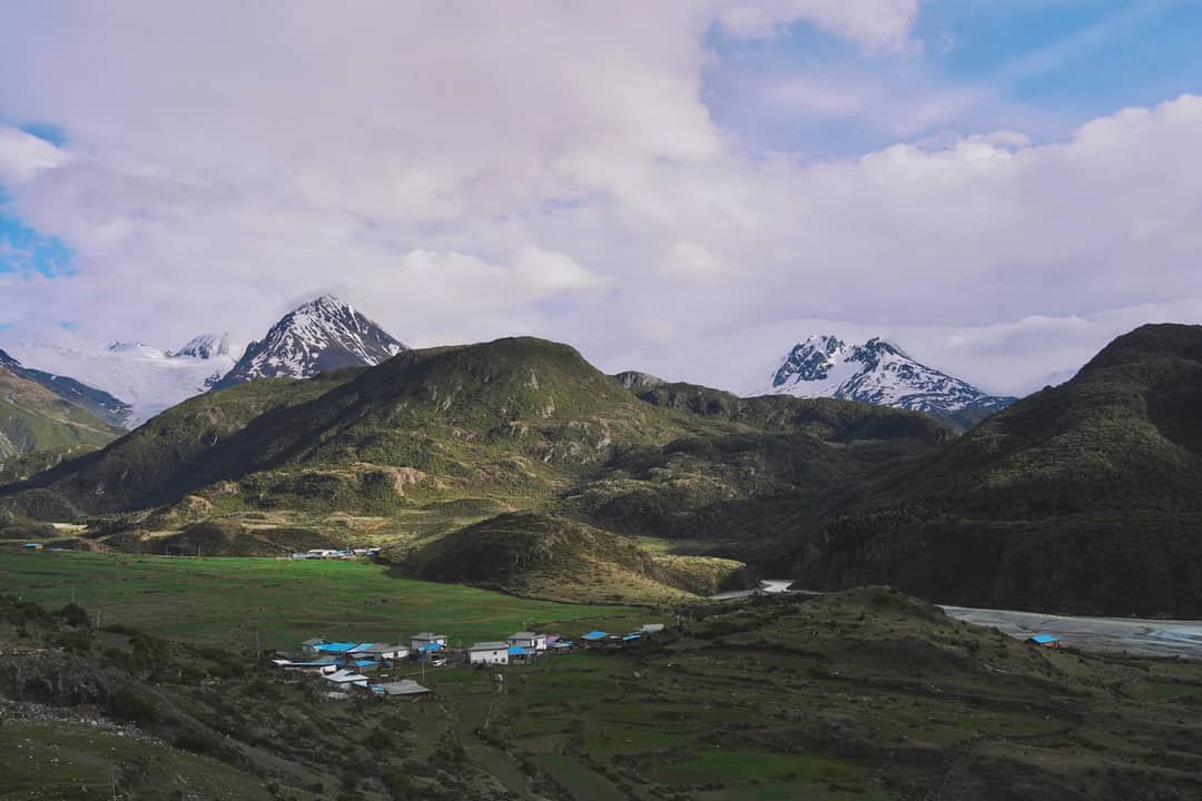 Picturesque village nestled in lush mountains with snow-capped peaks under a cloudy sky.