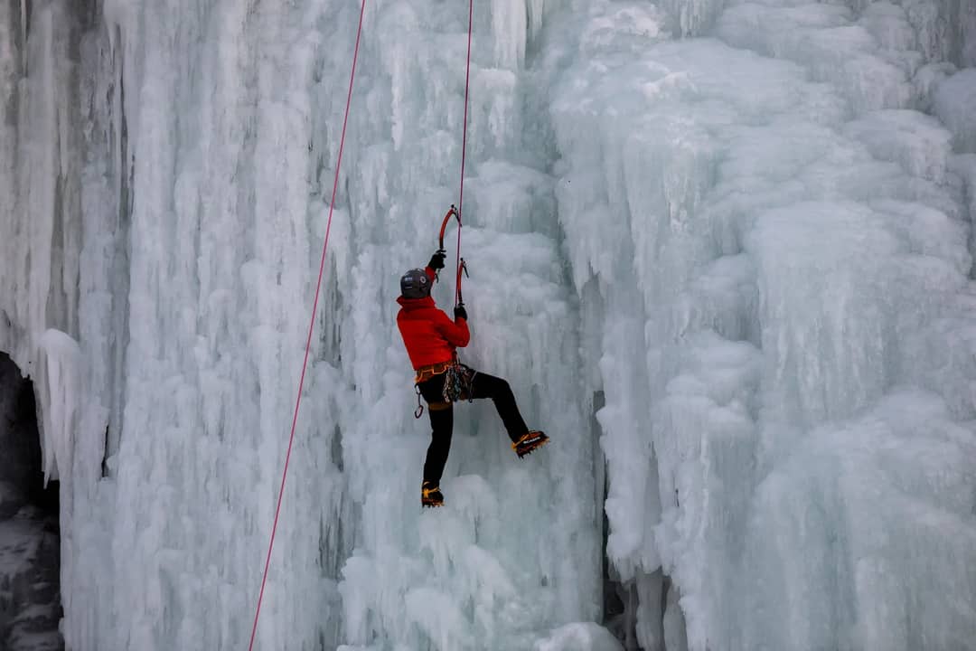 A climber in red jacket ascends a massive frozen waterfall, showcasing their ice climbing skills.