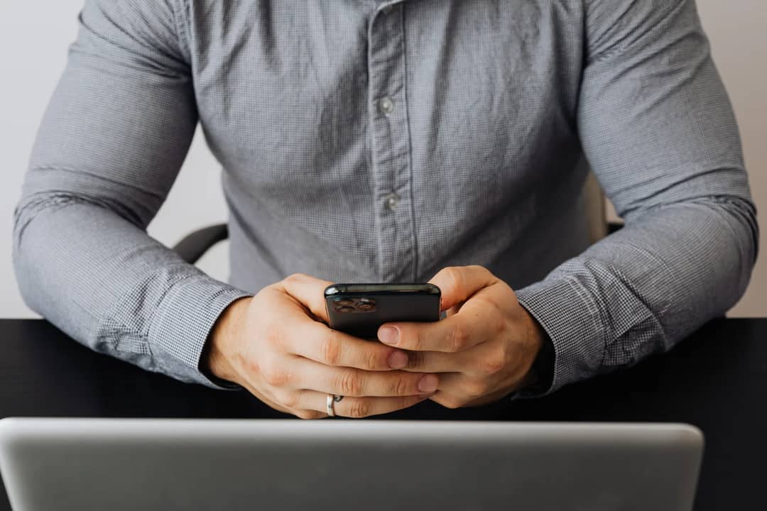 A person wearing a gray shirt holds a smartphone in front of a laptop, possibly at work.