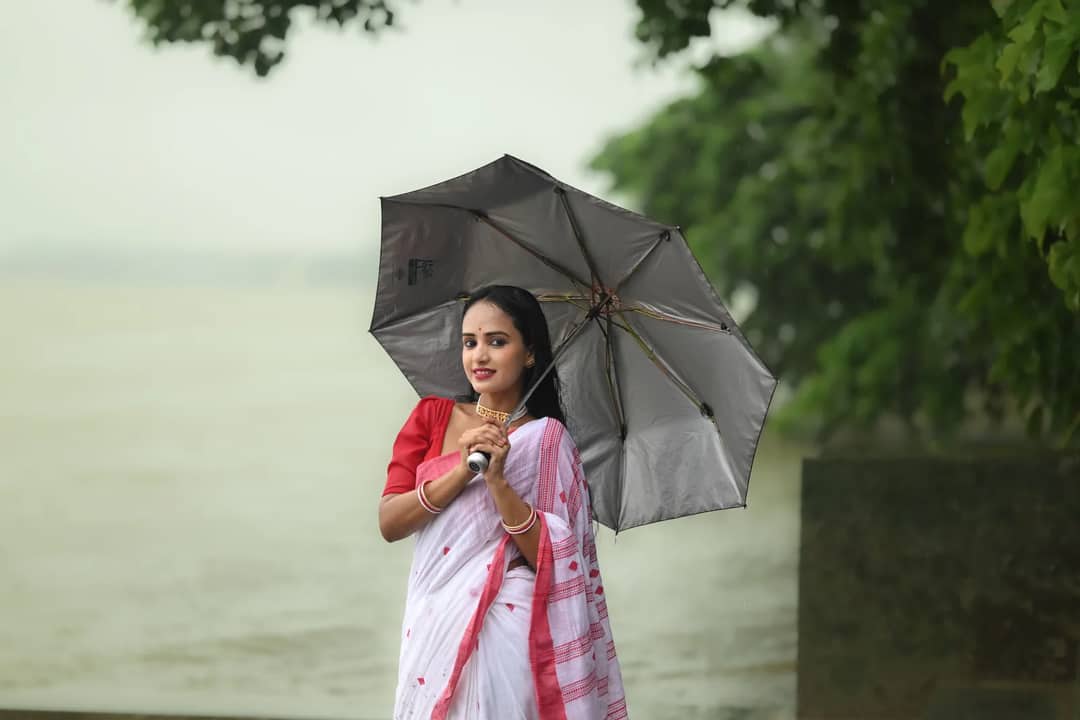 A woman in traditional attire holding an umbrella by a riverside, surrounded by lush greenery.
