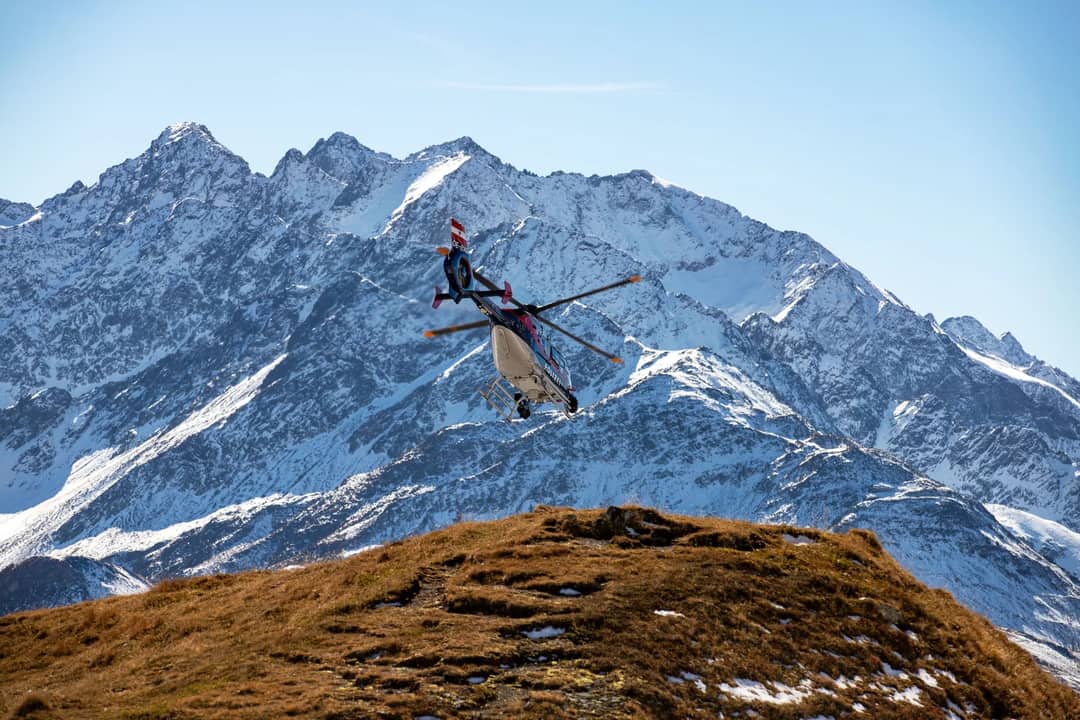 A helicopter flies over the snowy peaks of the Korutany mountains on a clear day.