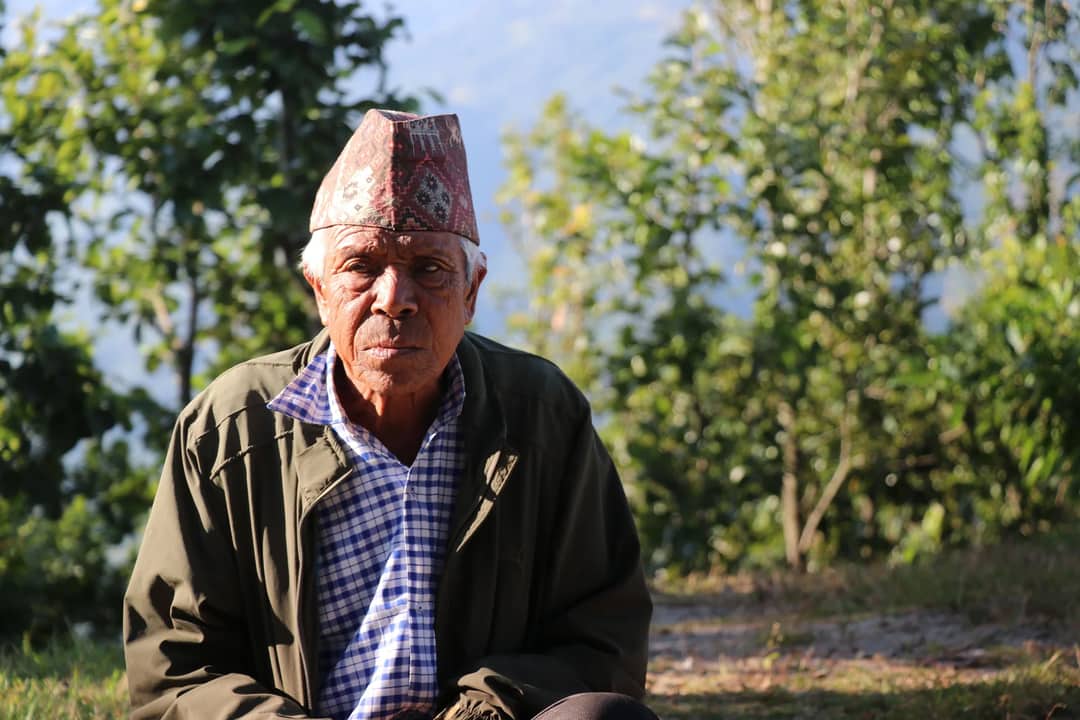 An elderly man wearing traditional Nepali headwear sits in a natural setting with trees.