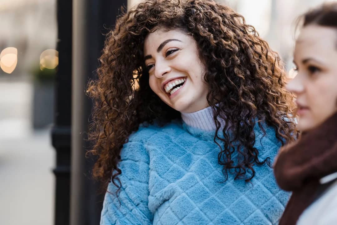 Cheerful young ethnic lady with long curly hair in warm sweater laughing an looking away while spending time on city street with female friend