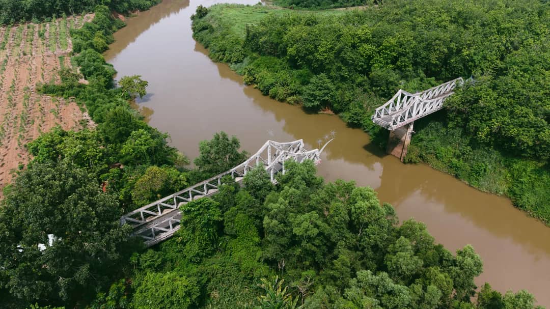 Aerial shot of a broken bridge spanning a river amidst lush greenery and farmland.