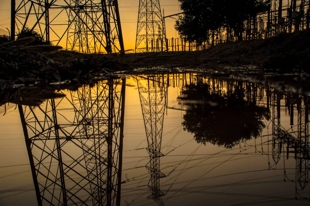 Silhouette of power towers reflecting in a puddle during sunset, creating an industrial scene.