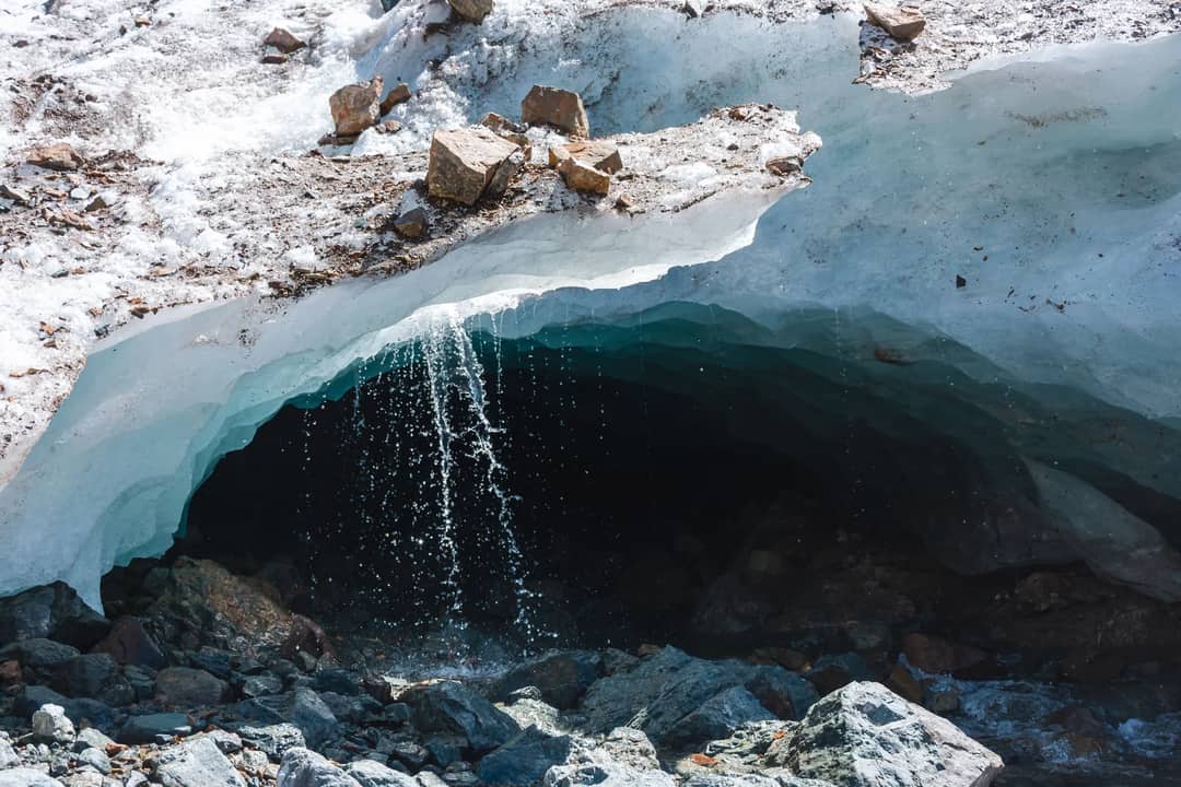 A picturesque view of a melting ice cave with water flowing, surrounded by snow and stones.