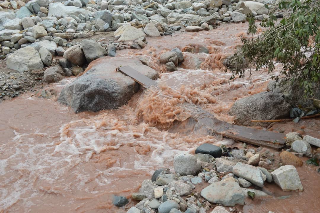 Muddy floodwater pouring over boulders and a broken plank crossing