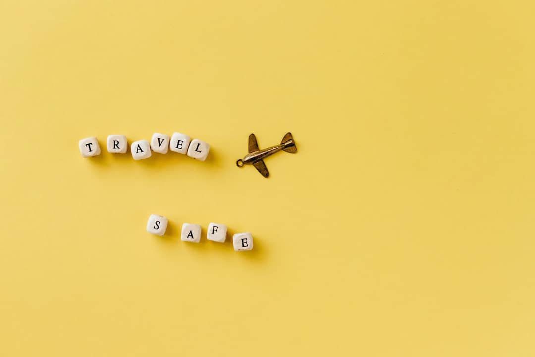 Flat lay of 'Travel Safe' text with airplane pendant on yellow background, evoking travel and safety concepts.