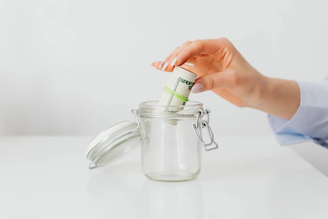 A close-up of a woman's hand putting rolled US dollar bills into a glass jar, symbolizing saving and budgeting.