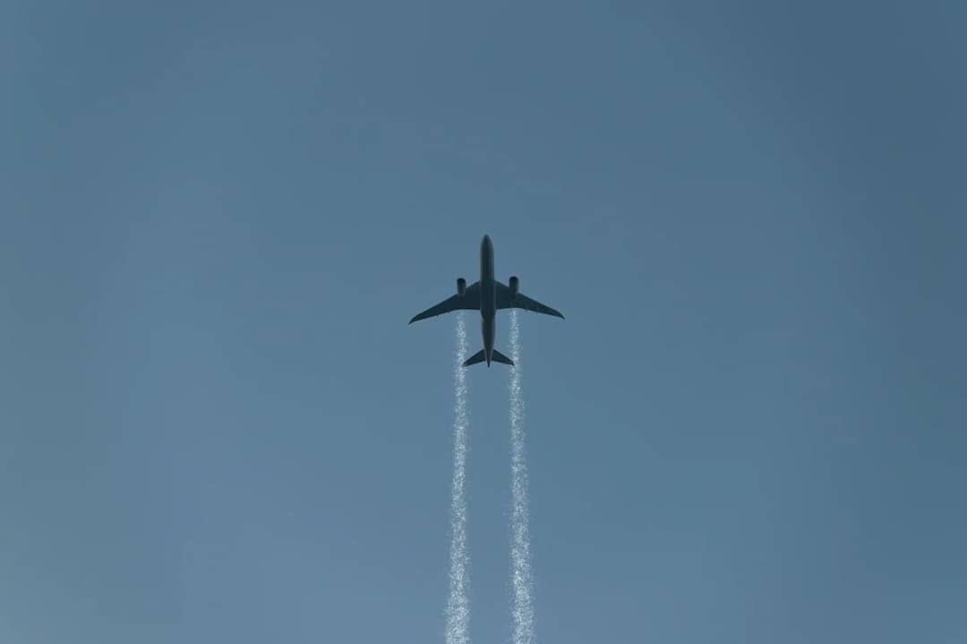 A commercial airplane leaving contrails as it flies across a clear blue sky, viewed from below.