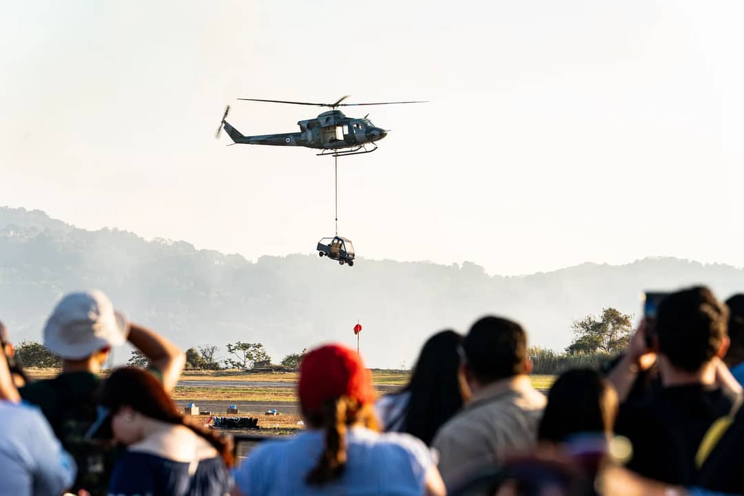 Crowd watches a helicopter air display lifting a vehicle in El Salvador.
