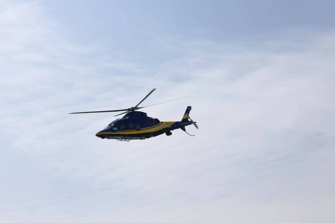 Blue and yellow helicopter flying across cloudy sky on a clear day, showcasing aviation in action.