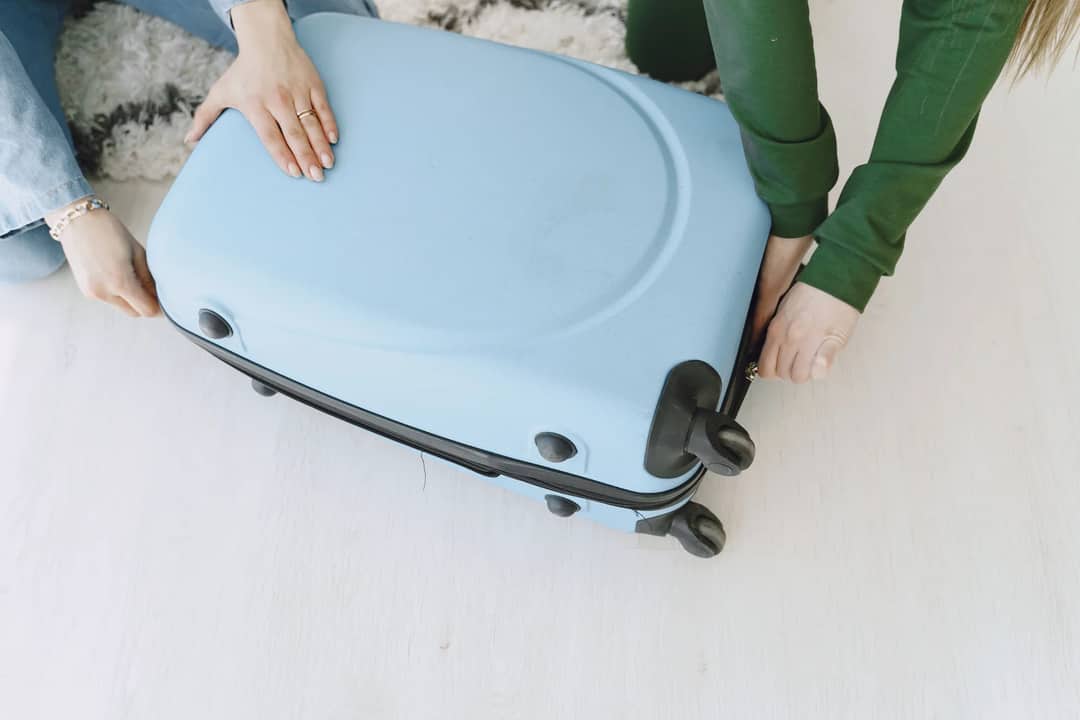 Unrecognizable female friends in casual clothes sitting on floor with fluffy carpet and opening suitcase together while preparing for journey and adventure