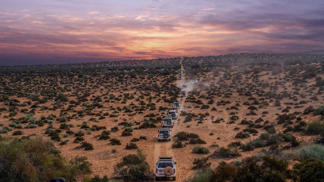 A convoy of vehicles travels through a vast desert landscape under a stunning sunset.
