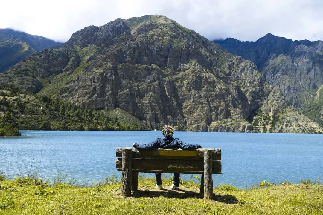 A person relaxes on a bench by Phoksundo Lake in Nepal amidst stunning mountain scenery.