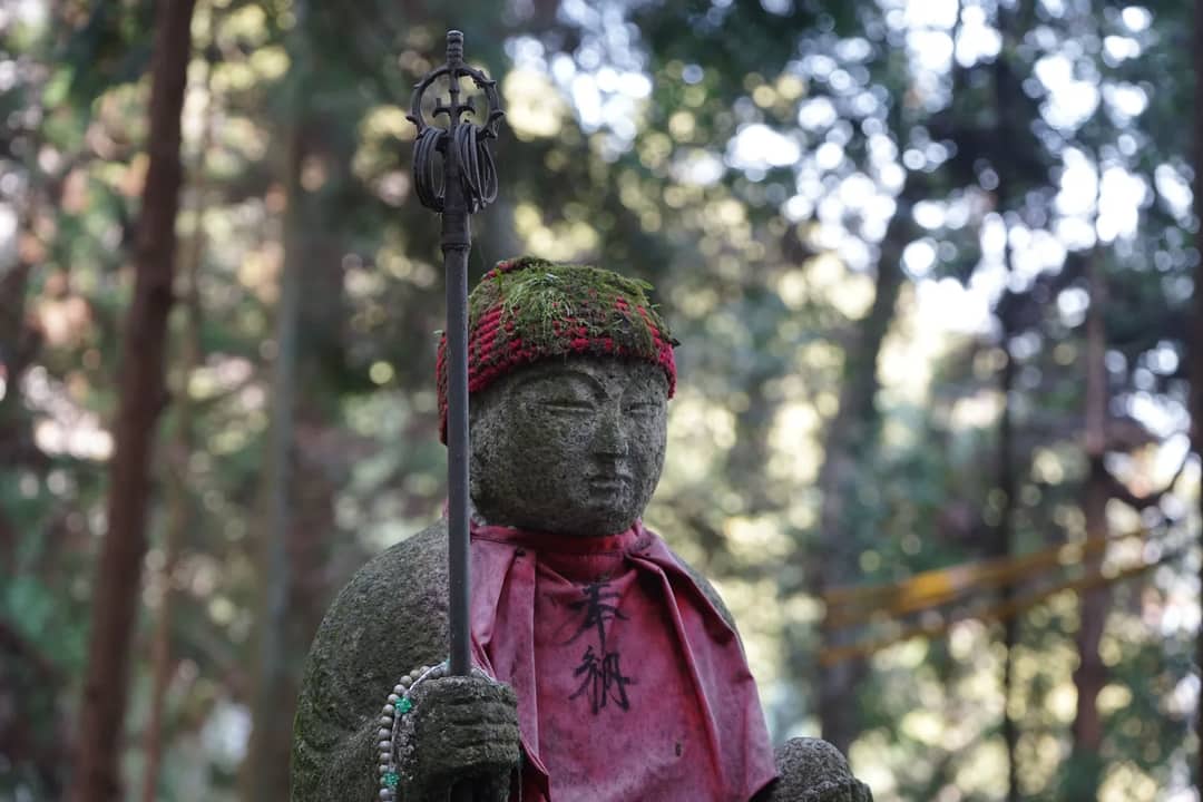 Moss-covered stone statue adorned with red cloth, set in a peaceful forest clearing.