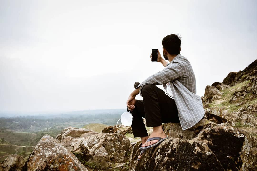 Man sits on rocky terrain capturing a selfie with scenic mountain background.