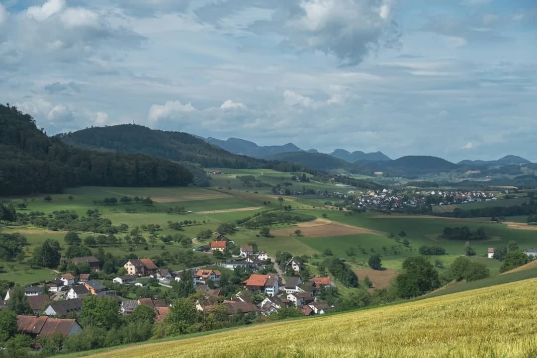 A serene rural landscape featuring lush fields, a small village, and distant mountains under a cloudy summer sky.