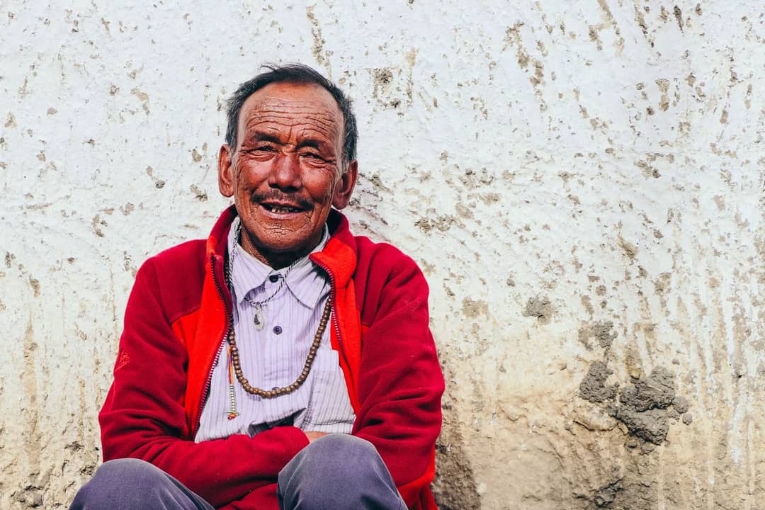 Elderly man in Hikkim, India, wearing a red jacket and necklace, smiling against a textured wall.