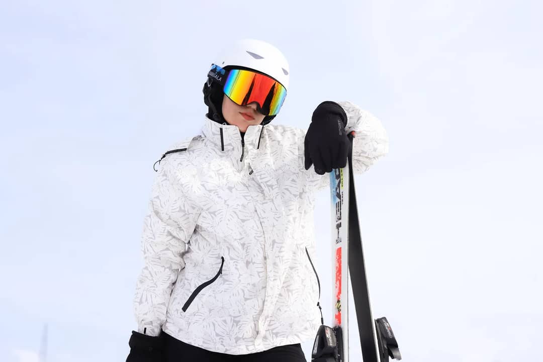 A woman wearing a ski helmet and goggles poses with skis against a snowy backdrop.