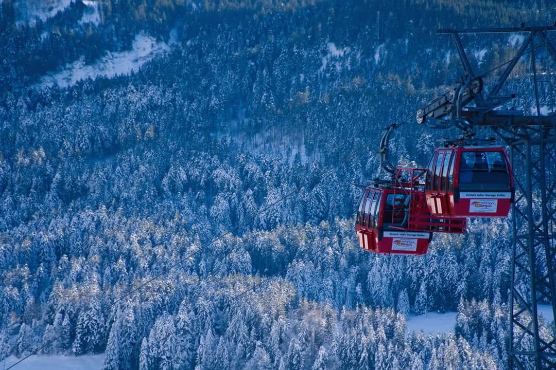 A red cable car travels over a snow-covered forest with mountain views.