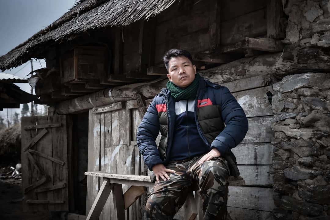 A man in a puffer jacket sits seriously outside a wooden house in rural Nepal.