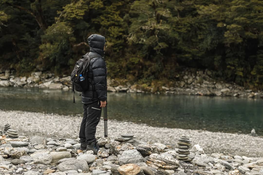 Hiker by a peaceful river, bundled for winter adventure in lush nature.