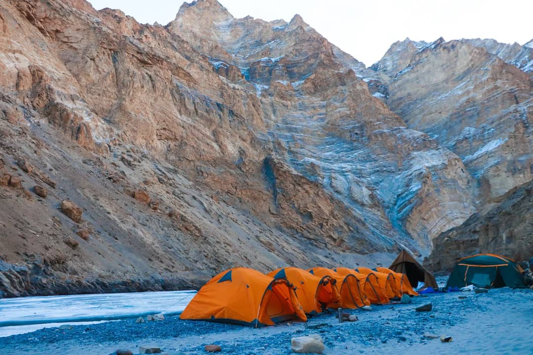 Row of orange tents on a rocky terrain against majestic mountains in Ladakh at daylight.