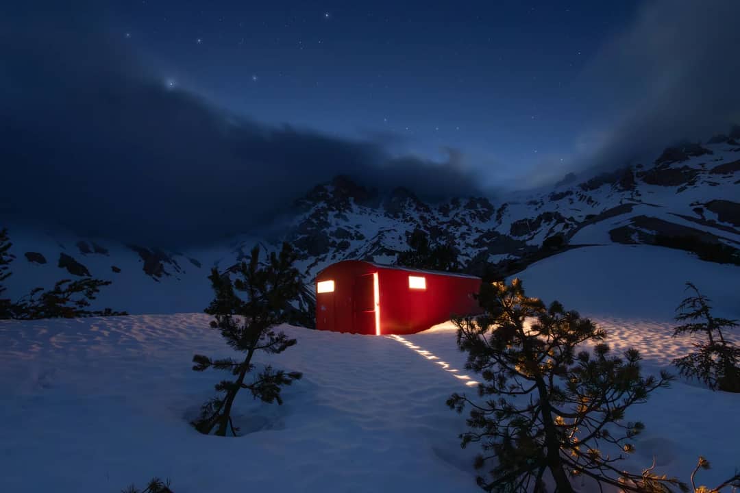 Red mountain hut illuminated under a starry night sky, surrounded by snow and trees.