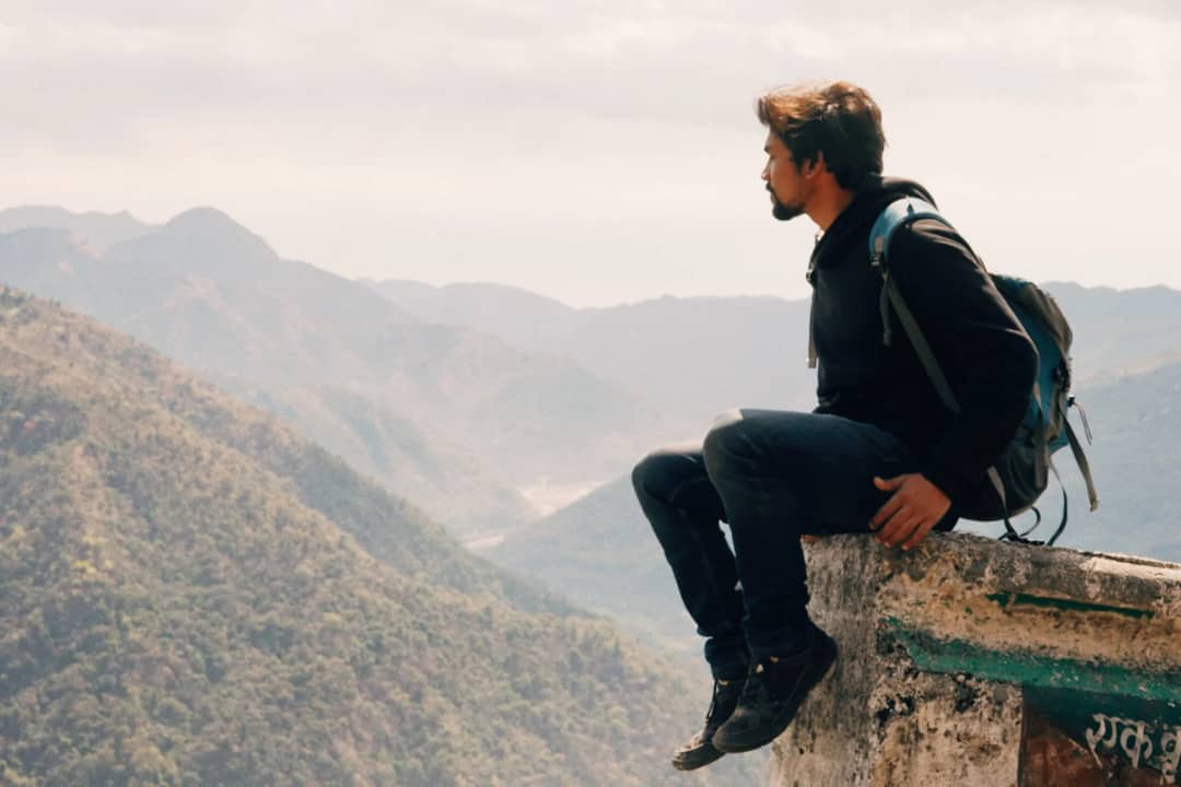 Side view of traveling man with backpack sitting on high edge with landscape of mountains in haze
