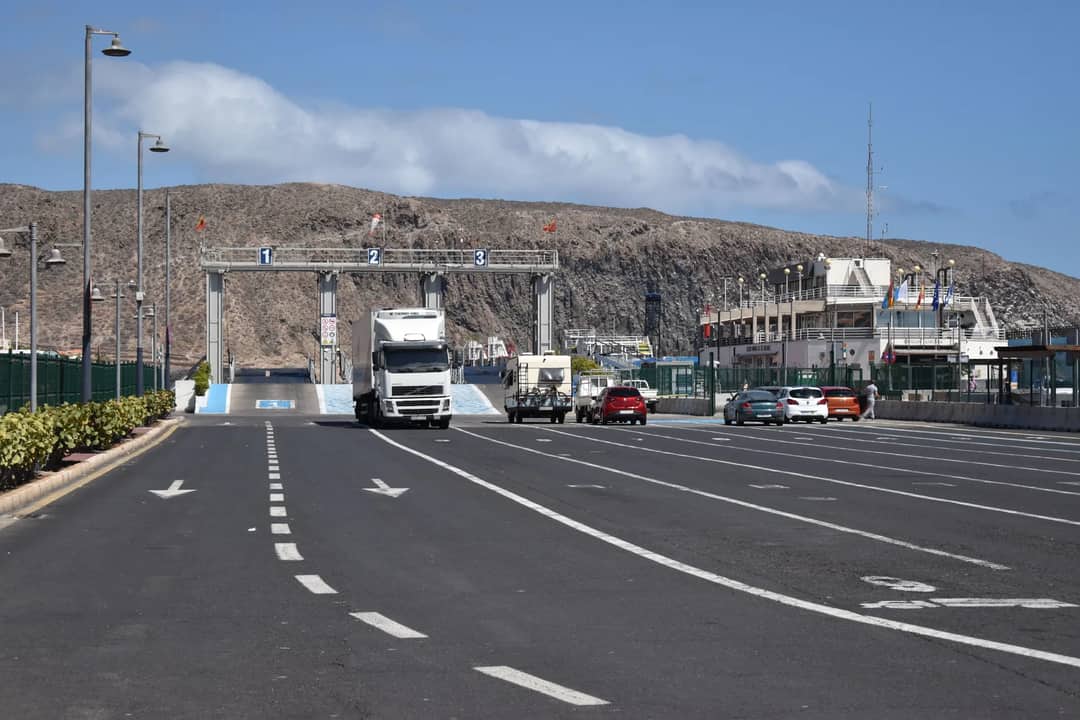 A ferry terminal with trucks and cars under a clear blue sky and dry hills in the background.