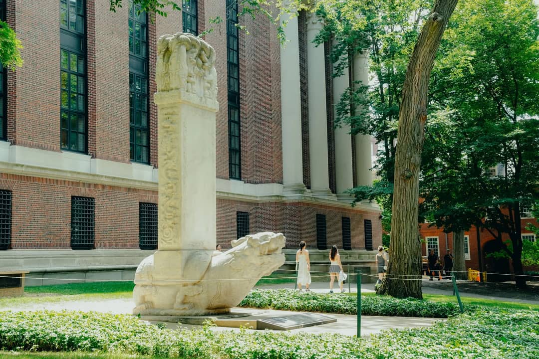 A historic stone sculpture near a red-brick building on a sunny day in Harvard Yard.