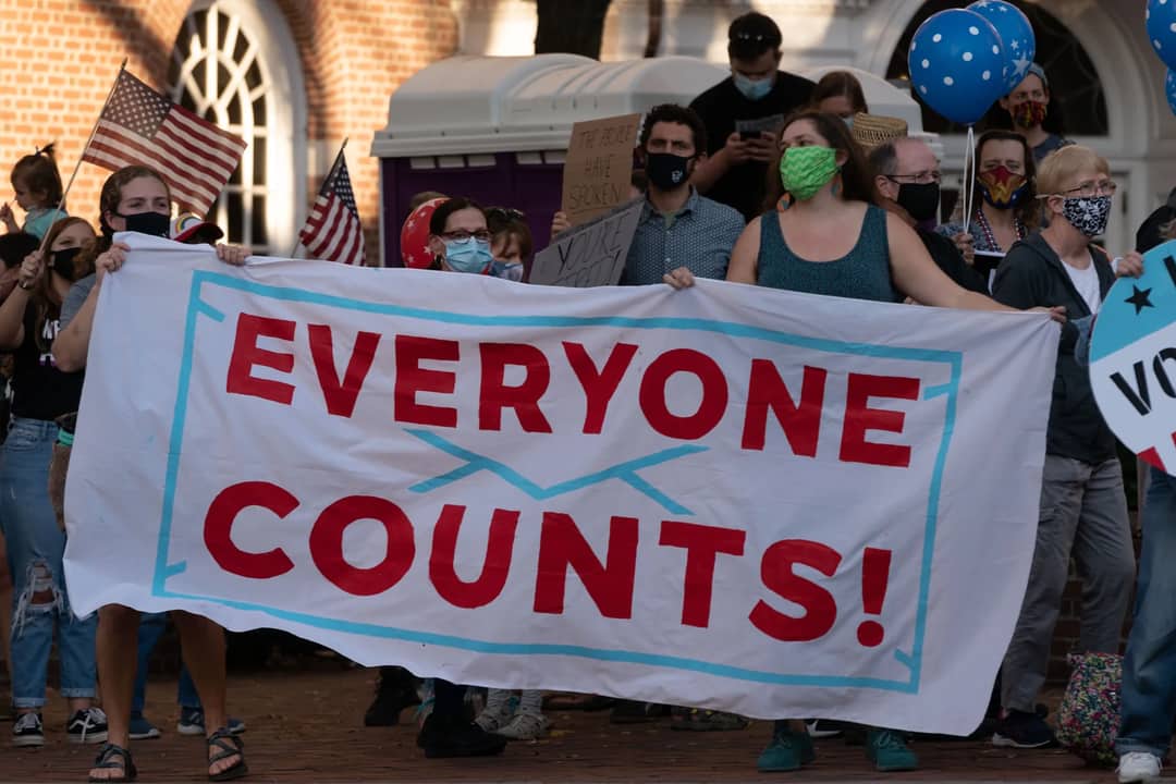 A diverse group of masked protestors holding a 'Everyone Counts' banner advocating for democratic rights.