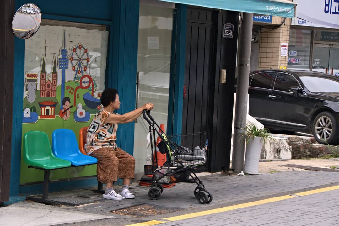 Elderly woman sits by a colorful mural on Daegu street, capturing urban life.