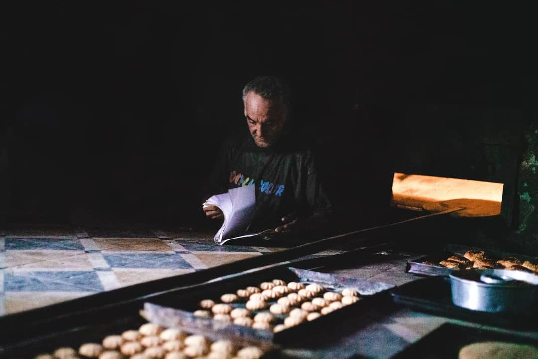 A traditional Moroccan baker reading a book surrounded by cookies and pastries in a dimly lit bakery.
