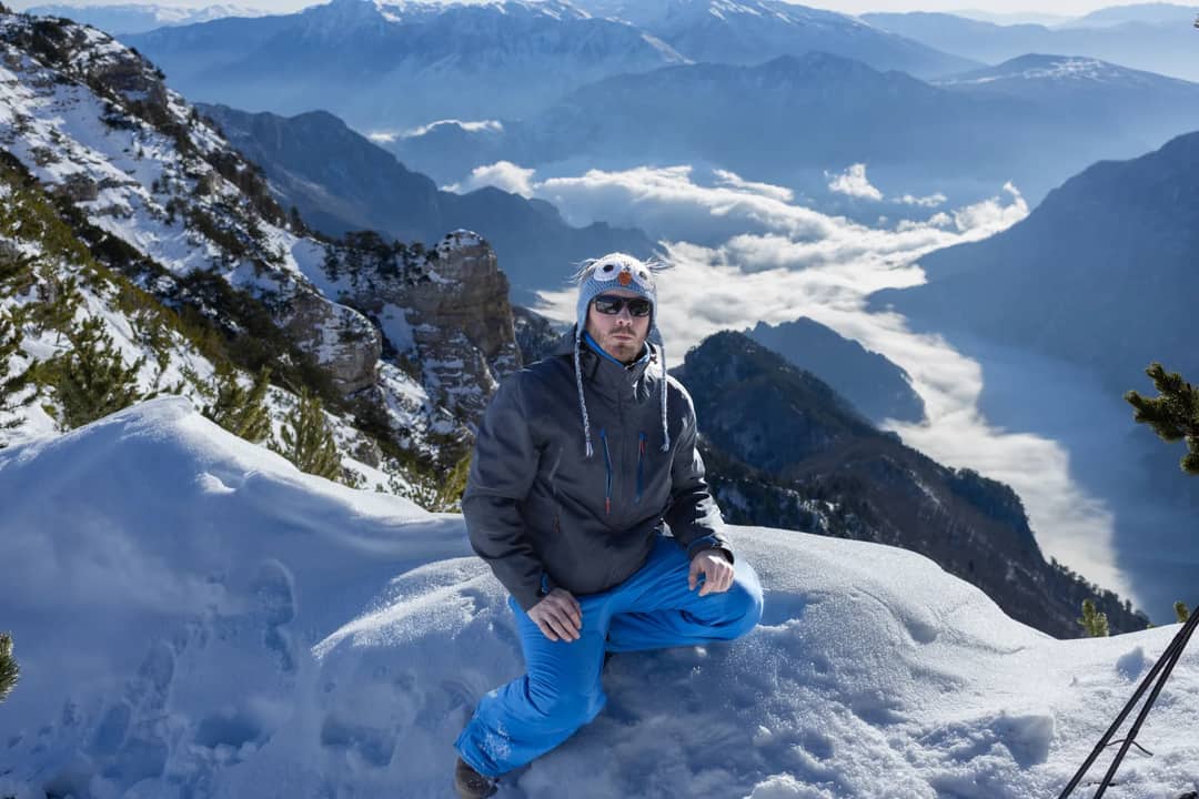 Male adventurer poses on a snowy mountain peak with breathtaking views of the sky and valleys.