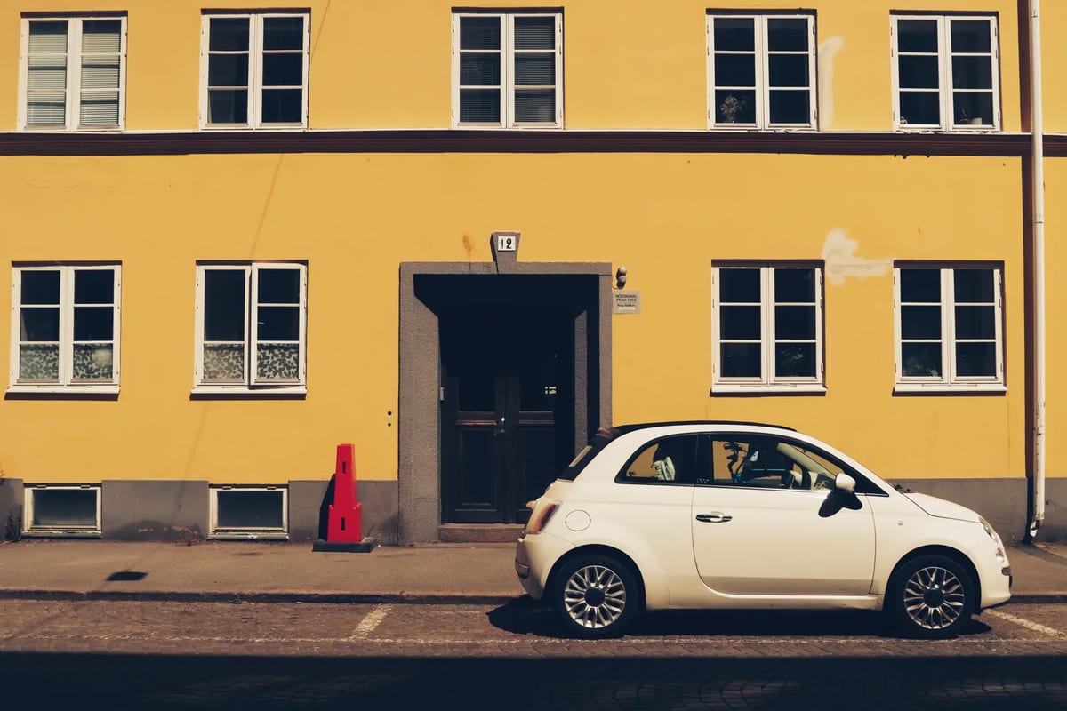A small white city car parked outside a yellow building