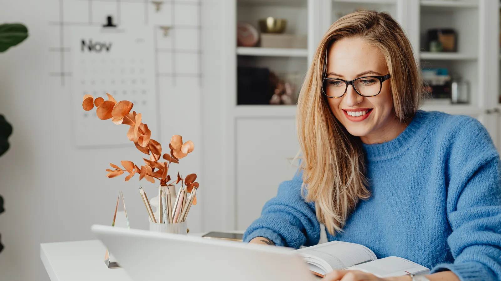 A smiling woman in glasses and a blue jumper works on a laptop at a white desk, a November wall calendar behind her