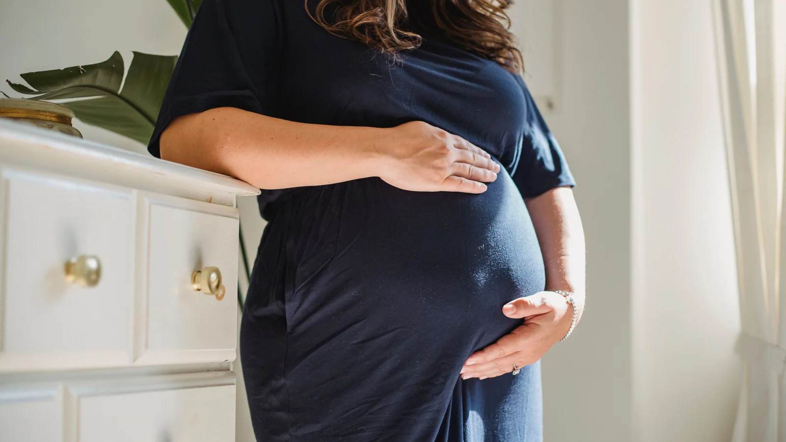Pregnant woman in a navy dress resting a hand on her bump in a bright room
