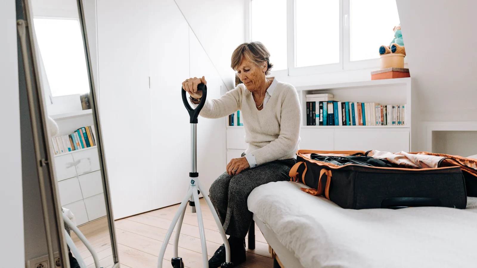An older woman sits on a bed beside an open suitcase in a bright white bedroom, one hand resting on a walking aid
