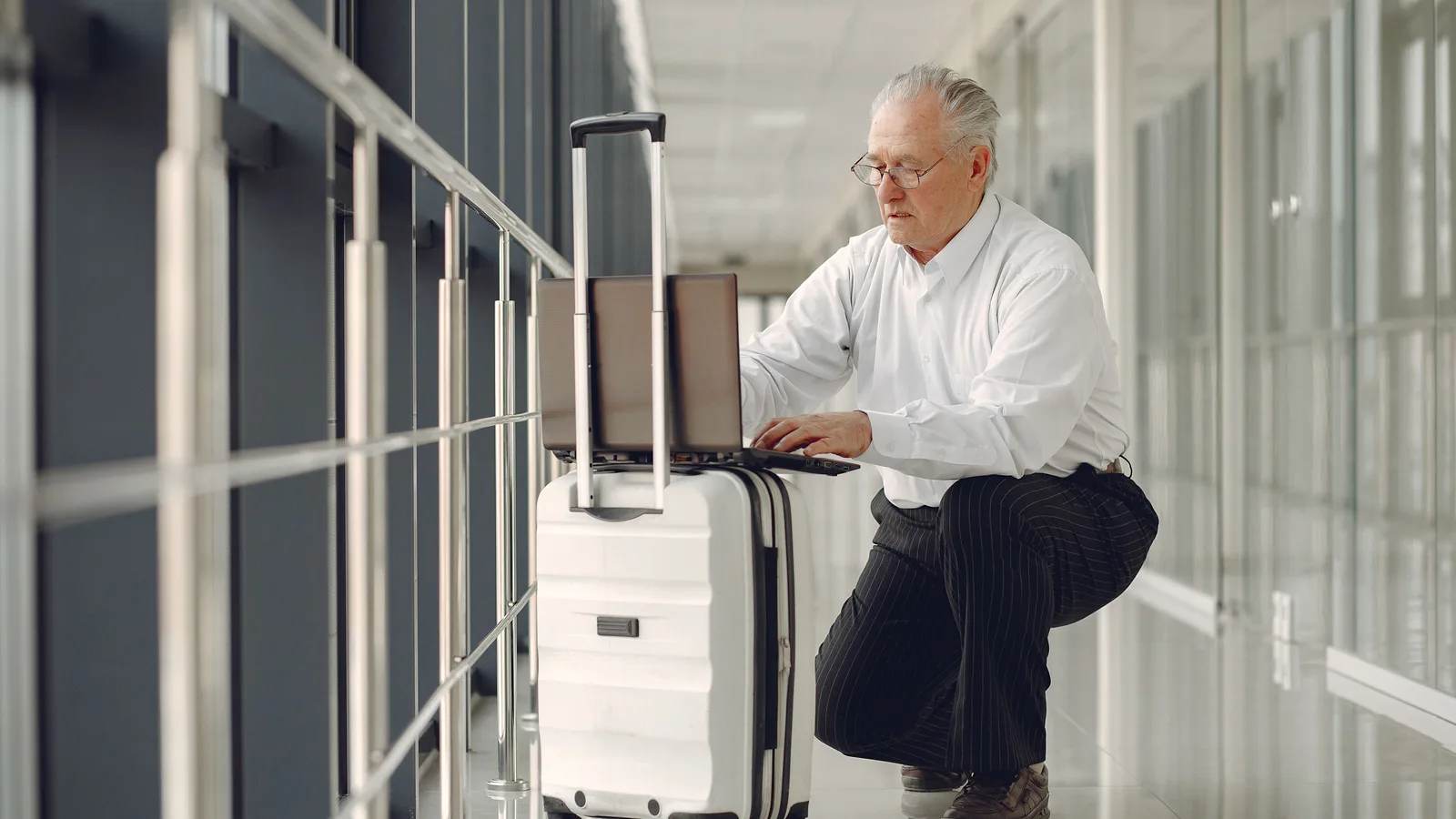 Older man with grey hair and glasses crouching beside a white suitcase in a bright airport corridor, typing on a laptop balanced on it