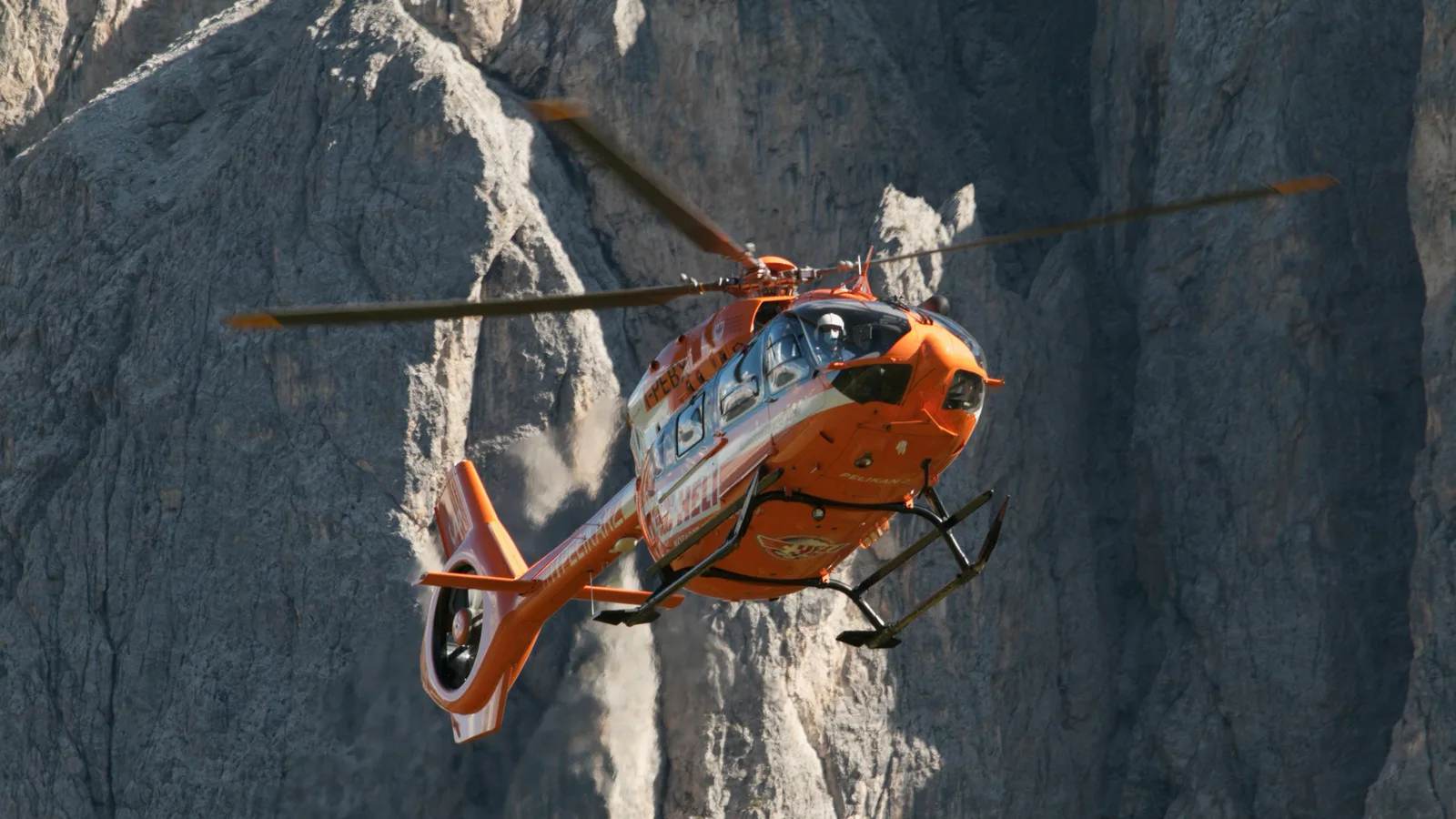 An orange rescue helicopter flying beside a mountain cliff
