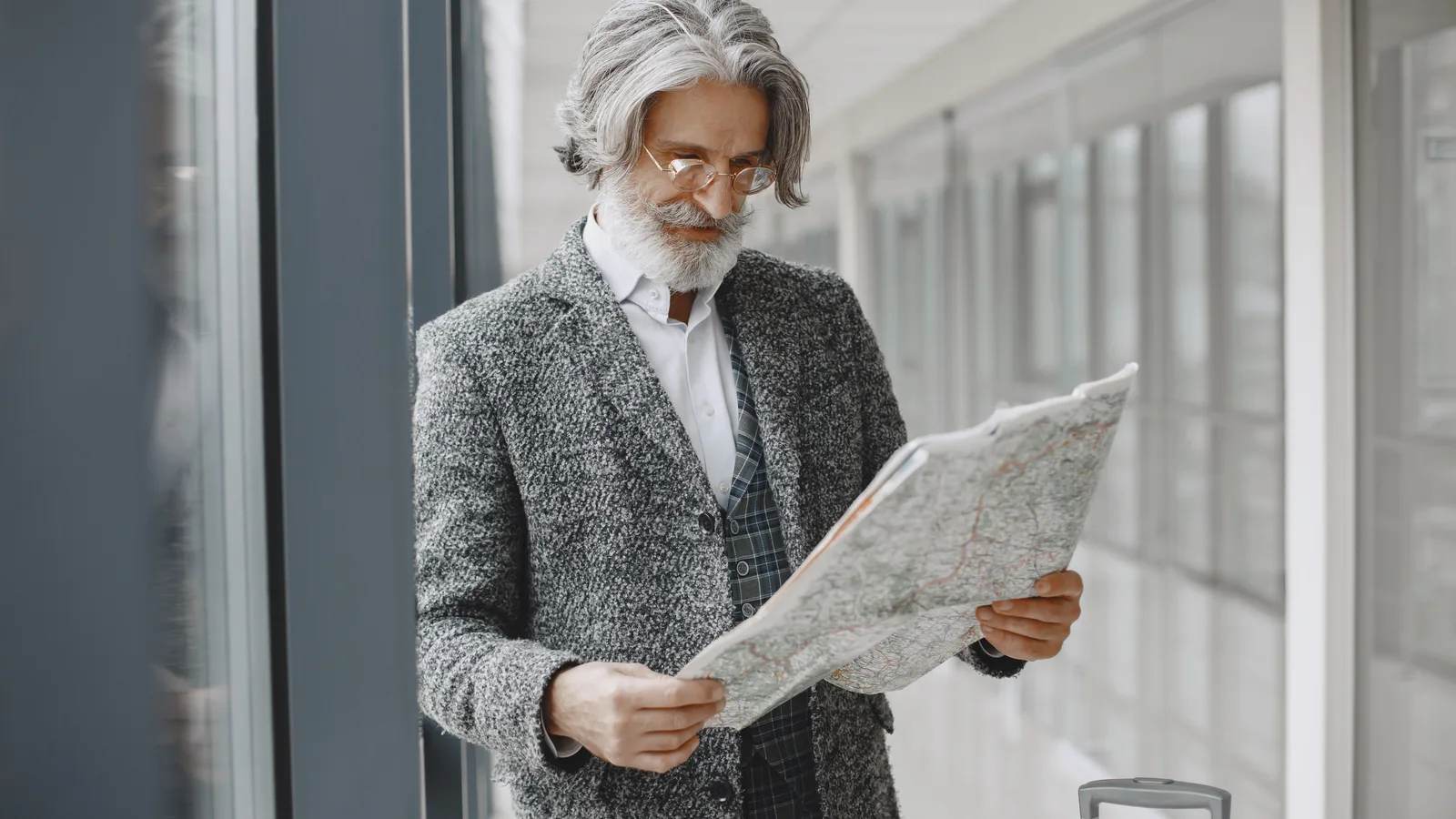 An older man with grey hair and glasses reads a paper map in a bright airport corridor, a suitcase handle beside him