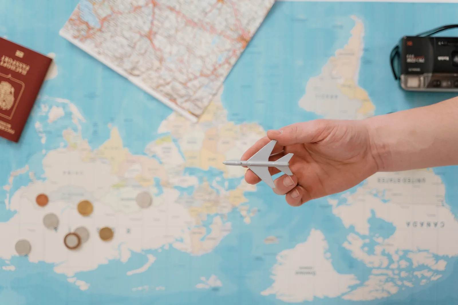 A hand holds a small toy aeroplane over a world map surrounded by coins, a camera and a passport — planning the budget for a year of travel.