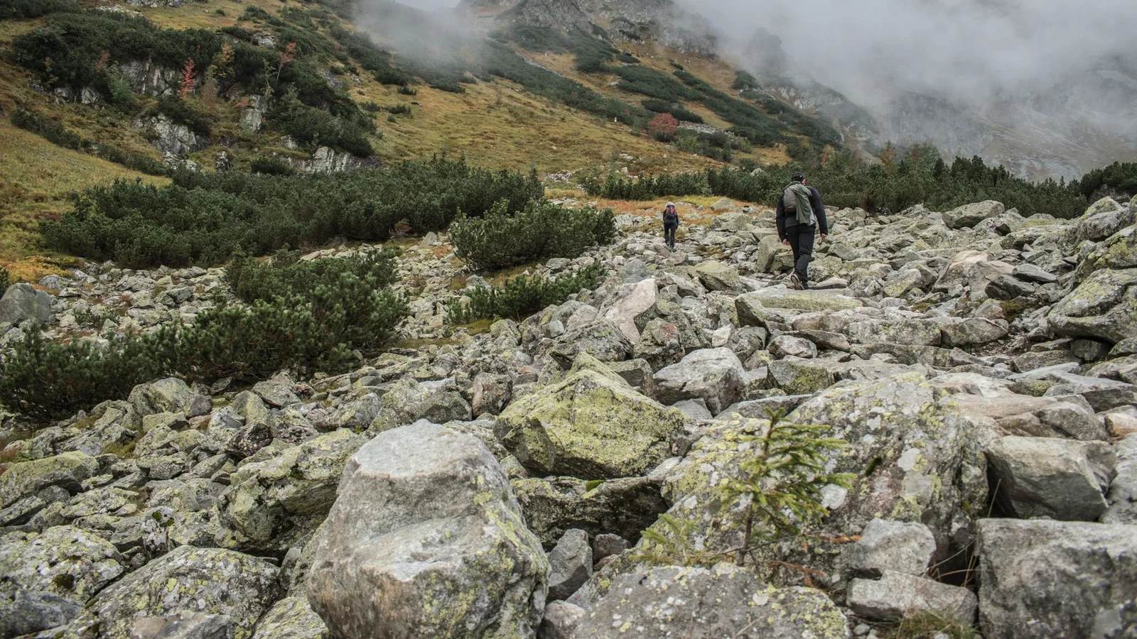Two walkers picking a line across a boulder field in low cloud — loose rock underfoot is the ground the whole boot-or-shoe argument is really about.